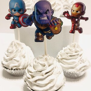 May include: Six white cupcakes with different superhero character toppers. The toppers are Captain America, Thanos, Iron Man, Hulk, Thor, and Spiderman.
