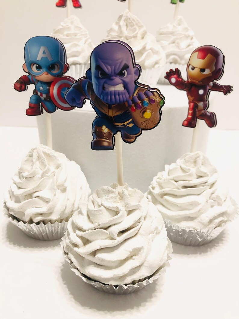 May include: Six white cupcakes with different superhero character toppers. The toppers are Captain America, Thanos, Iron Man, Hulk, Thor, and Spiderman.