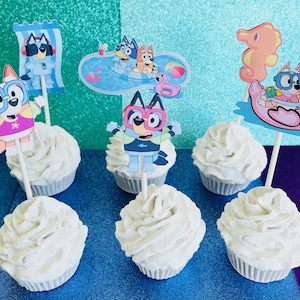 May include: White frosted cupcakes with character toppers featuring a cartoon dog in swim attire. The toppers include a dog on a beach towel, in a pool, and riding a flamingo float. The cupcakes are on a blue and teal background.