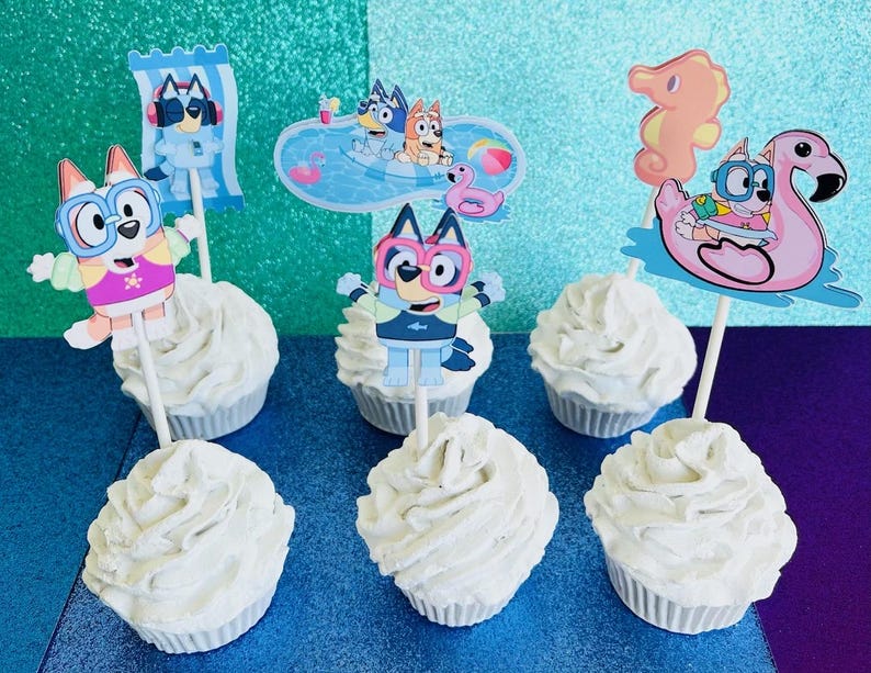 May include: White frosted cupcakes with character toppers featuring a cartoon dog in swim attire. The toppers include a dog on a beach towel, in a pool, and riding a flamingo float. The cupcakes are on a blue and teal background.