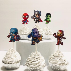 May include: Six white cupcakes with different superhero character toppers. The toppers include Spider-Man, Thor, Hulk, Captain America, Thanos, and Iron Man.