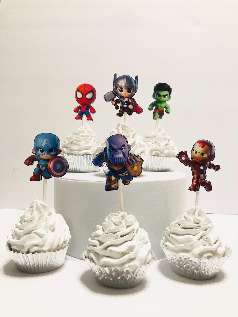 May include: Six white cupcakes with different superhero character toppers. The toppers include Spider-Man, Thor, Hulk, Captain America, Thanos, and Iron Man.