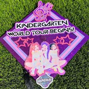 May include: A purple glitter graduation cap with the text "KINDERGARTEN WORLD TOUR BEGINS!" and a musical note design. The cap features a graphic of three cartoon figures and pink star accents.