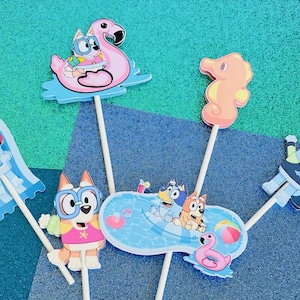 May include: A collection of colorful Bluey-themed cake toppers. The toppers feature Bluey and friends in various summer-themed designs, including a flamingo float, seahorse, and a pool scene. Each topper is attached to a wooden stick.