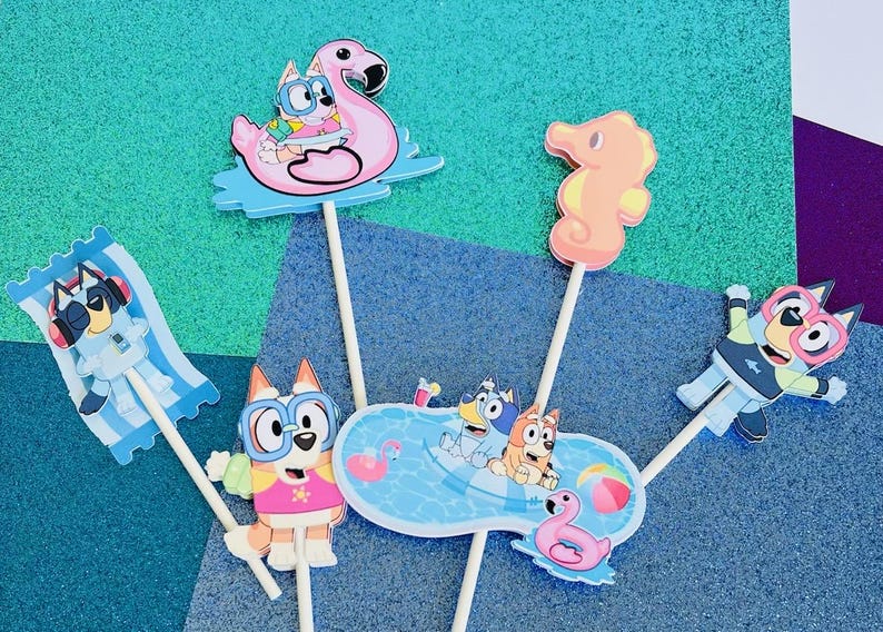 May include: A collection of colorful Bluey-themed cake toppers. The toppers feature Bluey and friends in various summer-themed designs, including a flamingo float, seahorse, and a pool scene. Each topper is attached to a wooden stick.