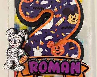 May include: A layered cake topper featuring the number two with a purple background patterned with Halloween icons like ghosts, bats, and candy. A mummy character stands to the left, and a banner at the bottom displays the name ROMAN in purple.