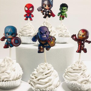 May include: Set of 7 cupcake toppers featuring various superhero characters, including Captain America, Iron Man, Hulk, Thor, Spiderman, and Thanos. The toppers are printed on cardstock and attached to wooden sticks. The cupcakes are frosted with white frosting.