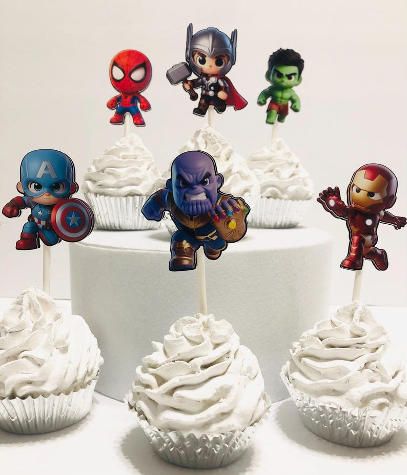 May include: Set of 7 cupcake toppers featuring various superhero characters, including Captain America, Iron Man, Hulk, Thor, Spiderman, and Thanos. The toppers are printed on cardstock and attached to wooden sticks. The cupcakes are frosted with white frosting.