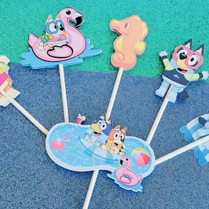 May include: Colorful cartoon character cake toppers featuring a flamingo, seahorse, and pool scene. The toppers depict characters in sunglasses and swimwear, perfect for a summer-themed party. The toppers are attached to wooden sticks.