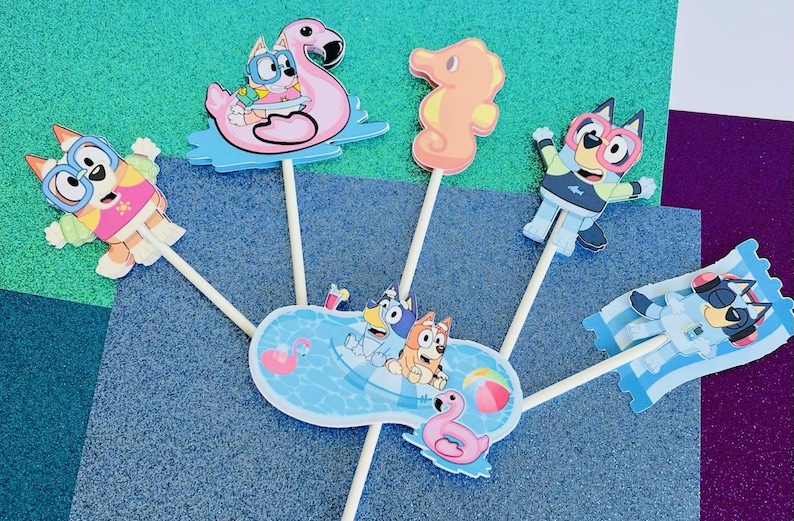 May include: Colorful cartoon character cake toppers featuring a flamingo, seahorse, and pool scene. The toppers depict characters in sunglasses and swimwear, perfect for a summer-themed party. The toppers are attached to wooden sticks.