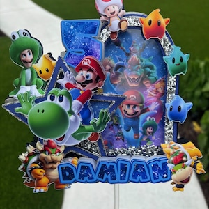 K&ouml;nnte beinhalten: Ein mehrschichtiger Tortenaufleger mit einer gro&szlig;en blauen Zahl 5 und dem Namen DAMIAN in blauen Buchstaben. Das Design enth&auml;lt verschiedene bunte Charakter-Ausschnitte aus der Super Mario-Reihe, silberne Glitzerakzente und eine zentrale Szene.