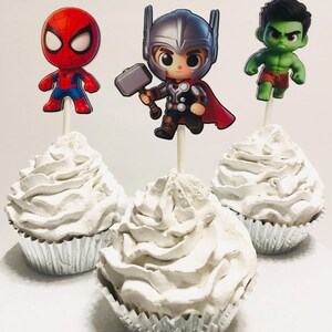 May include: Three cupcakes decorated with white frosting and topped with superhero character cake toppers. The toppers are Spiderman, Thor, and the Hulk.