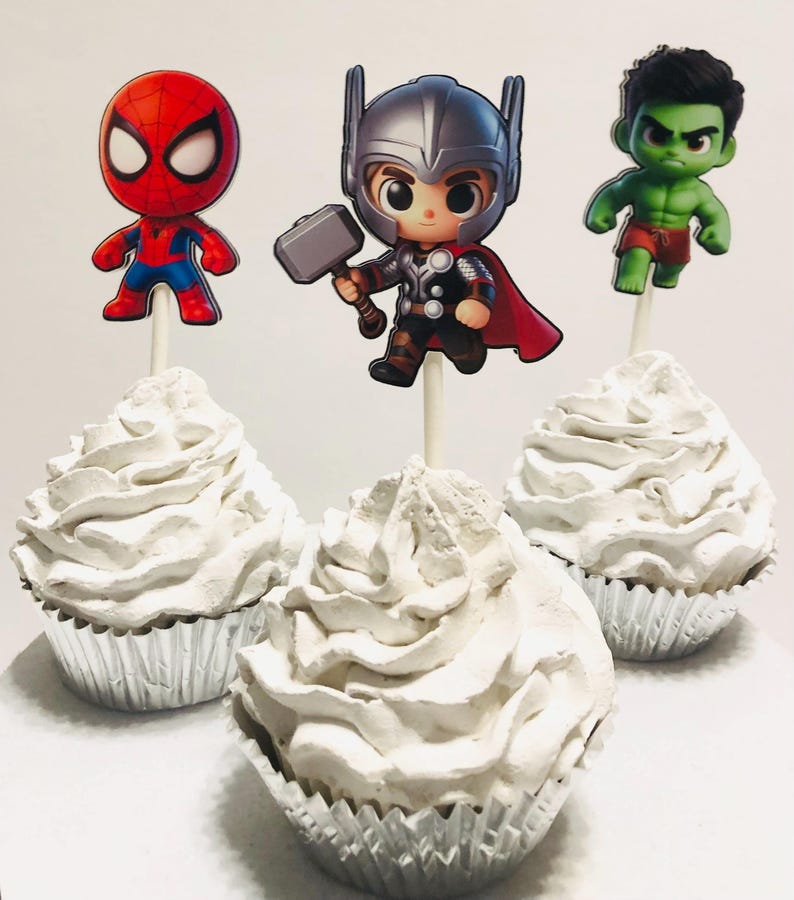 May include: Three cupcakes decorated with white frosting and topped with superhero character cake toppers. The toppers are Spiderman, Thor, and the Hulk.