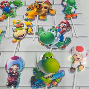 以下が含まれることがあります： マリオ、ヨッシー、クッパ、クッパJr.、キノピオのカラフルなキャラクターカットアウトを特徴とするスーパーマリオをテーマにしたカップケーキトッパーのコレクション。各カットアウトにはきらめくホログラフィックの縁取りがあり、細い白いプラスチックの棒に取り付けられています。