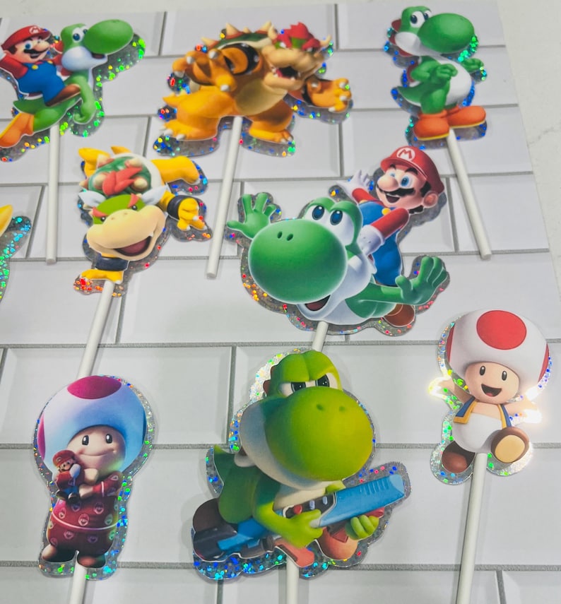 以下が含まれることがあります： マリオ、ヨッシー、クッパ、クッパJr.、キノピオのカラフルなキャラクターカットアウトを特徴とするスーパーマリオをテーマにしたカップケーキトッパーのコレクション。各カットアウトにはきらめくホログラフィックの縁取りがあり、細い白いプラスチックの棒に取り付けられています。