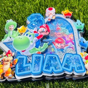 K&ouml;nnte beinhalten: Ein mehrschichtiger Tortenaufleger im Super Mario-Design mit dem Namen LIAM in blauen Glitzerbuchstaben. Das Design enth&auml;lt Ausschnitte von Mario, Yoshi, Bowser und Toad sowie ein zentrales Sch&uuml;ttelfenster mit silbernen Pailletten und Bildern.