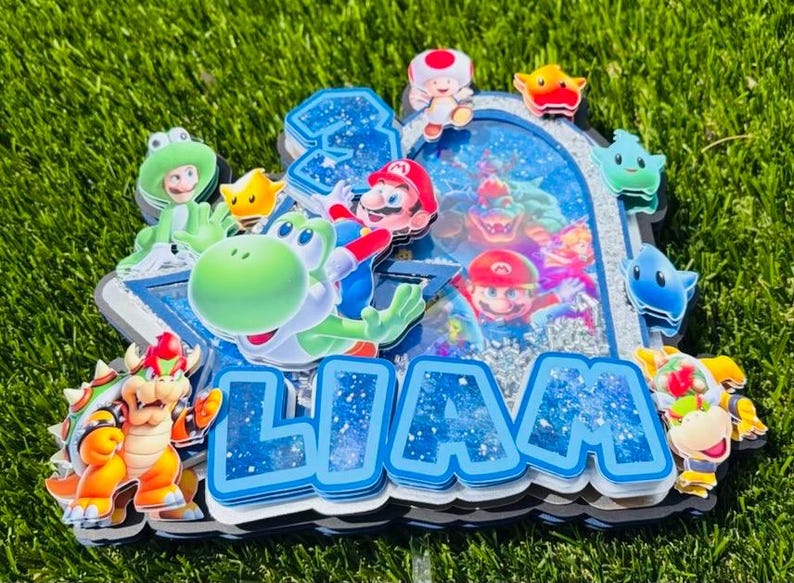 K&ouml;nnte beinhalten: Ein mehrschichtiger Tortenaufleger im Super Mario-Design mit dem Namen LIAM in blauen Glitzerbuchstaben. Das Design enth&auml;lt Ausschnitte von Mario, Yoshi, Bowser und Toad sowie ein zentrales Sch&uuml;ttelfenster mit silbernen Pailletten und Bildern.