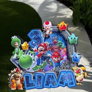 K&ouml;nnte beinhalten: Ein mehrschichtiger Tortenaufleger im Super Mario-Design mit dem Namen LIAM in blauen Buchstaben mit Galaxienmuster. Das Design enth&auml;lt Ausschnitte von Mario, Yoshi, Bowser, Toad und verschiedenen Sternen sowie ein zentrales Element mit silbernem Glitzer.