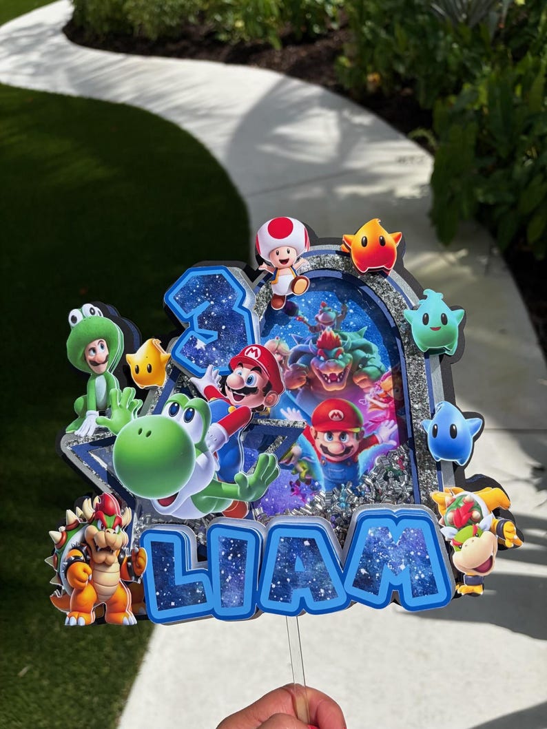 K&ouml;nnte beinhalten: Ein mehrschichtiger Tortenaufleger im Super Mario-Design mit dem Namen LIAM in blauen Buchstaben mit Galaxienmuster. Das Design enth&auml;lt Ausschnitte von Mario, Yoshi, Bowser, Toad und verschiedenen Sternen sowie ein zentrales Element mit silbernem Glitzer.
