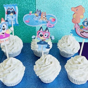 May include: Six cupcakes with white frosting, each topped with a character from the animated series Bluey. The toppers feature Bluey and Bingo in swimwear, sunglasses, and pool floats. The background is a sparkly teal and purple.