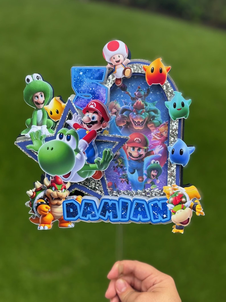 K&ouml;nnte beinhalten: Ein mehrschichtiger Tortenaufleger mit dem Namen DAMIAN und der Zahl 5. Das Design zeigt verschiedene Super Mario-Figuren wie Mario, Yoshi, Bowser und Toad vor einem glitzernden blau-silbernen Hintergrund mit Sternenmotiven.