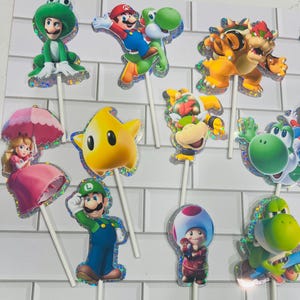 以下が含まれることがあります： マリオ、ルイージ、ピーチ姫、クッパ、クッパJr.、キノピオ、ヨッシーなど、スーパーマリオのキャラクターが描かれたカップケーキトッパーのセット。各切り抜きにはホログラムの縁取りがあり、装飾用の白いスティックに取り付けられています。