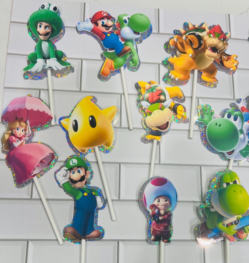 以下が含まれることがあります： マリオ、ルイージ、ピーチ姫、クッパ、クッパJr.、キノピオ、ヨッシーなど、スーパーマリオのキャラクターが描かれたカップケーキトッパーのセット。各切り抜きにはホログラムの縁取りがあり、装飾用の白いスティックに取り付けられています。