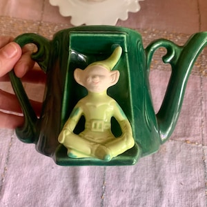 May include: Green ceramic teapot with a sculpted elf figure in a sitting position. The elf is wearing a green and yellow outfit and has a white face.