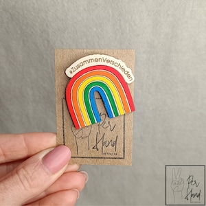 Könnte beinhalten: Eine hölzerne Regenbogen-Anstecknadel mit roten, orangen, gelben, grünen und blauen Streifen. Der obere Teil des Regenbogens ist gewölbt und mit dem schwarzen Text #ZusammenVerschieden bedruckt. Die Nadel ist auf einer braunen Pappkarte befestigt.