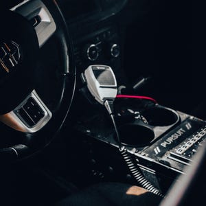 May include: Interior view of a vehicle's dashboard, featuring a steering wheel, a white microphone, and a control panel. The control panel has the text "PURSUIT //" and several buttons. The overall color scheme is dark, with silver accents.