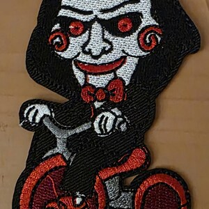 Op de afbeelding: Geborduurde patch met een personage met een wit gezicht, rode ogen en een rode vlinderdas, die op een rode driewieler rijdt. Het personage draagt een zwarte cape. De patch is ontworpen met zwart, rood, wit en grijs garen.