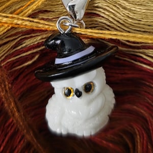 Könnte beinhalten: Ein weißer Eulenanhänger mit einem schwarzen Hexenhut und einem silbernen Verschluss. Die Eule hat goldfarbene Augen und ist vor einem Hintergrund aus farbenfrohem Garn platziert. Ein skurriles Accessoire für Halloween oder ein Fantasy-Thema.