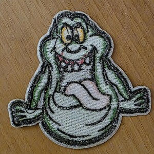 Puede incluir: Parche bordado con un personaje de fantasma de dibujos animados con una amplia sonrisa y una larga lengua. El parche tiene un cuerpo verde claro, contorno negro y ojos amarillos. La lengua es rosa y los dientes son rojos. Para ropa o accesorios.