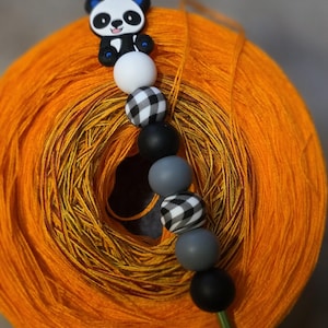 Puede incluir: Un ganchillo con un mango decorativo con cuentas que presenta un panda, cuentas negras, blancas, grises y de cuadros de búfalo, apoyado en una bola de hilo naranja vibrante. El gancho es verde.