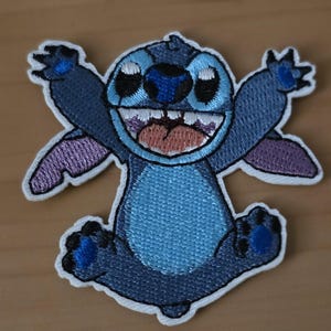 Könnte beinhalten: Gestickter Aufnäher mit der Figur Stitch aus Lilo & Stitch. Stitch wird in einer fröhlichen Pose mit ausgestreckten Armen und Beinen gezeigt. Das Design verwendet Blau-, Lila- und Weißtöne mit einem detaillierten Stickfinish. Der Aufnäher hat einen weißen Rand.