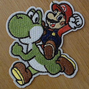 Könnte beinhalten: Gestickter Aufnäher mit Mario, der Yoshi reitet. Mario trägt einen roten Hut, blaue Latzhosen und einen Schnurrbart. Yoshi ist grün mit weißem Bauch und braunen Schuhen. Der Aufnäher hat einen weißen Rand, auf Holzuntergrund.