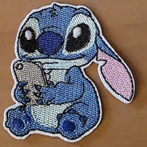 Könnte beinhalten: Gestickter Aufnäher mit der Figur Stitch aus Lilo & Stitch. Der blaue Außerirdische hält ein Telefon, mit großen schwarzen Augen, einem rosa Ohr und einem weißen Rand. Der Aufnäher ist für das Aufnähen auf Kleidung oder Accessoires gedacht.
