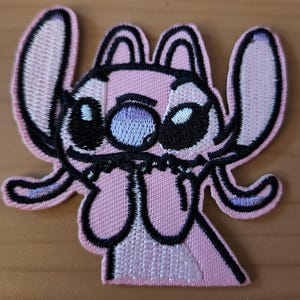 Könnte beinhalten: Rosa bestickter Aufnäher mit einer Cartoonfigur mit großen Augen, einem breiten Mund und großen Ohren. Die Figur ist schwarz umrandet, mit hellvioletten Akzenten in den Augen und Ohren. Für Kleidung oder Accessoires.