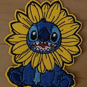 Peut inclure: Écusson brodé représentant un personnage de dessin animé bleu avec un grand sourire et des yeux noirs, entouré d'un tournesol jaune. Le patch a une bordure noire et est posé sur un fond neutre.