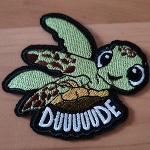 May include: Embroidered patch featuring a cartoon sea turtle with a smiling face and large eyes. The turtle is green with brown markings and black outlines. The word "DUUUUDE" is embroidered in white on a black background.