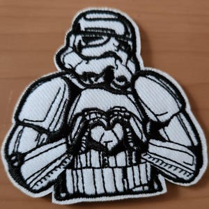Könnte beinhalten: Gestickter Aufnäher mit einem Stormtrooper aus Star Wars. Der schwarz-weiße Aufnäher zeigt den Helm und die Rüstung der Figur, wobei die Hände ein Herz formen. Der Aufnäher befindet sich auf einer hellbraunen Oberfläche.
