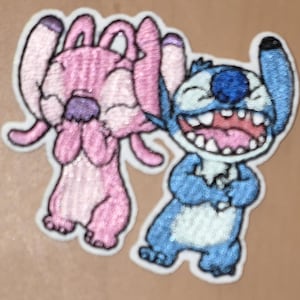 Könnte beinhalten: Gestickter Aufnäher mit zwei Zeichentrickfiguren, einer rosa und einer blauen. Die rosa Figur bedeckt ihr Gesicht, während die blaue Figur mit offenem Mund lacht. Beide haben weiße Umrisse.