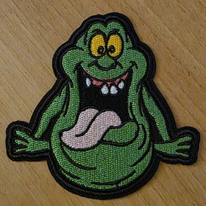 May include: Embroidered patch featuring a green cartoon ghost with yellow eyes, a pink tongue, and a wide, toothy grin. The patch has a black border and is likely for application on clothing or accessories.