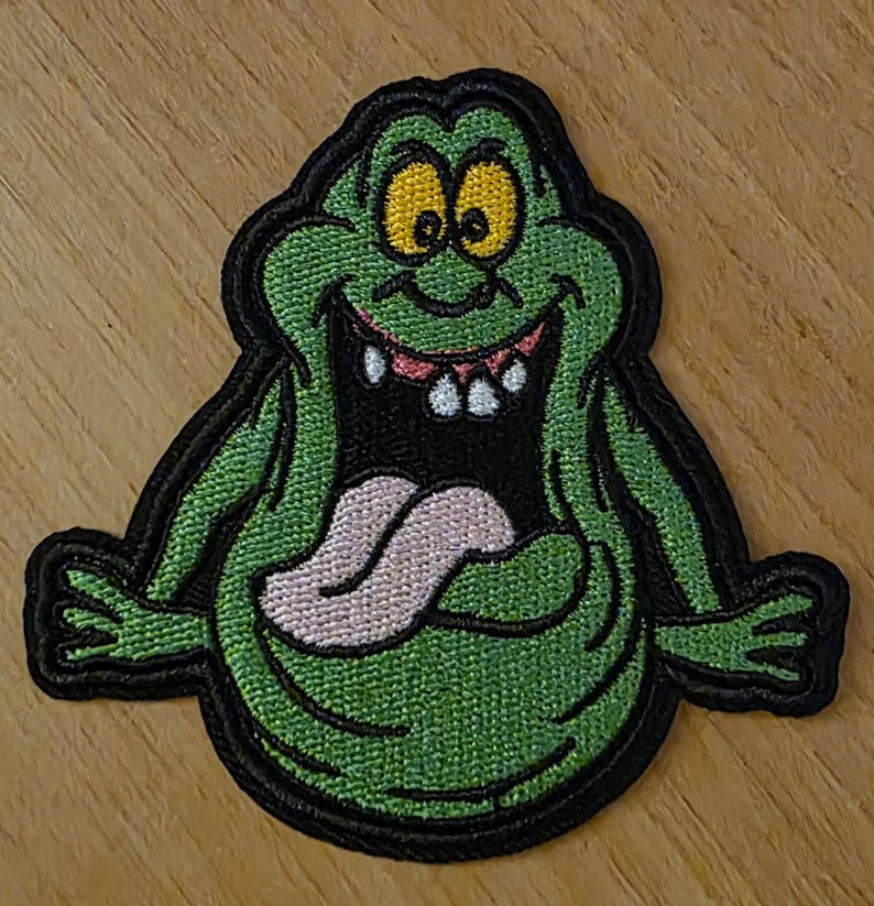 Puede incluir: Parche bordado que representa un fantasma de dibujos animados verde con ojos amarillos, una lengua rosa y una amplia sonrisa. El parche tiene un borde negro y est&aacute; dise&ntilde;ado para aplicarse en ropa o accesorios.
