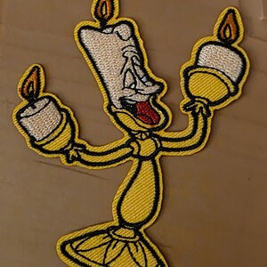 Könnte beinhalten: Gestickter Aufnäher mit einem gelben, anthropomorphen Kerzenleuchter-Charakter mit überraschtem Gesichtsausdruck und roter Zunge. Die Figur hat drei Kerzenflammen und ist schwarz umrandet. Der Aufnäher ist für Kleidung oder Accessoires.