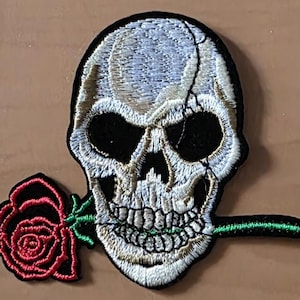 Könnte beinhalten: Gestickter Aufnäher mit einem Totenkopf, der eine Rose im Mund hält. Der Totenkopf ist weiß und schwarz, mit detaillierten Nähten. Die Rose ist rot mit grünen Blättern und Stiel. Der Aufnäher hat einen schwarzen Rand, für Kleidung oder Accessoires.
