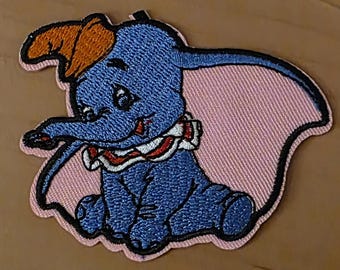 Peut inclure: Écusson brodé représentant Dumbo l'éléphant bleu avec un chapeau orange et un col à volants. L'écusson a un fond rose et une bordure noire. Le design est de style dessin animé.