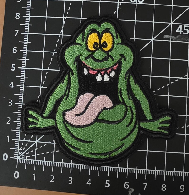 Puede incluir: Parche bordado con un fantasma verde de dibujos animados con ojos amarillos, una lengua rosa y una amplia sonrisa. El parche tiene un borde negro y est&aacute; sobre un fondo de cuadr&iacute;cula. El fantasma es un personaje de la franquicia Ghostbusters.