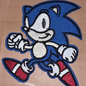Könnte beinhalten: Gestickter Aufnäher mit Sonic the Hedgehog in einer Laufpose. Der Aufnäher hat einen blauen Körper, weiße Handschuhe und einen weißen Bauch sowie rot-weiße Schuhe. Das Design ist von einem dunkelblauen Rand umgeben.