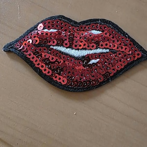 Könnte beinhalten: Ein roter Lippen-Aufnäher mit Pailletten und einem schwarzen Rand. Die Lippen sind schwarz umrandet und haben in der Mitte weiße Nähte. Dieser dekorative Aufnäher kann auf Kleidung oder Accessoires genäht oder aufgebügelt werden.